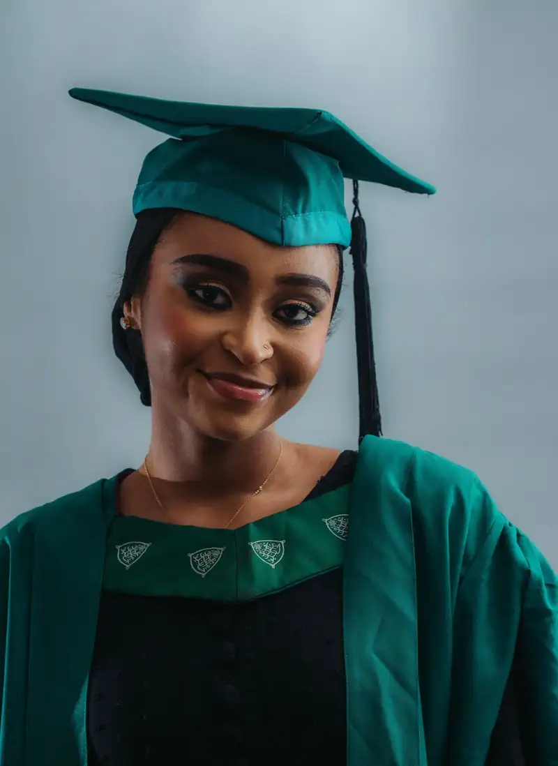Graduate in a black gown and red stole against a cool gray outdoor background. Stock photo from Pexels, not a Nafasi student.